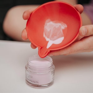 May include: A red funnel is held over a small glass jar filled with a light pink powder. The funnel has a small spout, and a small amount of the powder is visible inside. The jar has a clear label.