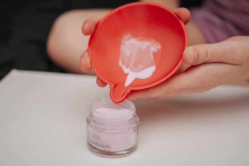 May include: A red funnel is held over a small glass jar filled with a light pink powder. The funnel has a small spout, and a small amount of the powder is visible inside. The jar has a clear label.