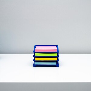 May include: A dark blue plastic desk organizer tray holding four stacks of square sticky notes in pink, lime green, yellow, and light blue. To the left sits a metallic blue planter containing a green and yellow variegated snake plant.