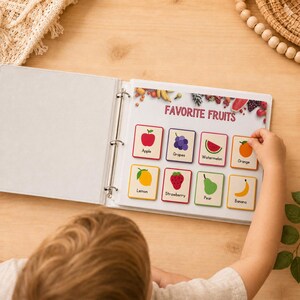 May include: A white binder open to a page titled "Favorite Fruits." The page displays colorful illustrations of various fruits, including an apple, grapes, watermelon, and orange. A child's hand is reaching for an orange card.