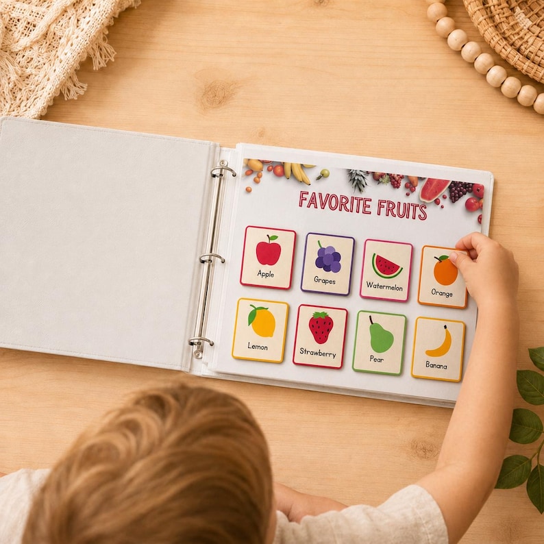 May include: A white binder open to a page titled "Favorite Fruits." The page displays colorful illustrations of various fruits, including an apple, grapes, watermelon, and orange. A child's hand is reaching for an orange card.