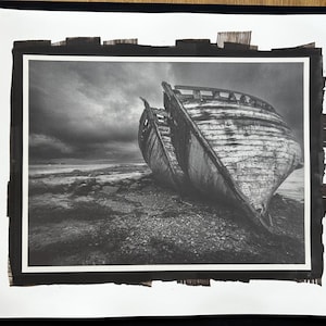 May include: Monochrome photograph depicting a wrecked boat on a stony beach beneath a dramatic, cloudy sky. The boat's wooden hull is broken and worn, with visible damage. The image conveys a feeling of abandonment and the effects of the elements.