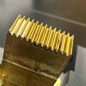 May include: Close-up of a gold-colored metal object with a ridged, comb-like edge. The object has a rectangular shape with a series of parallel grooves along one side. The metal has a slightly worn appearance, suggesting use or age.