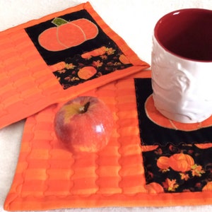 May include: Two orange quilted fabric coasters with a black fabric square featuring a pumpkin design. A white mug with a red interior is sitting on one of the coasters.