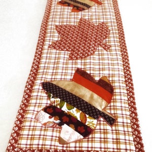 May include: A table runner with a plaid background and three appliqued fall leaves in shades of brown, red, and gold.