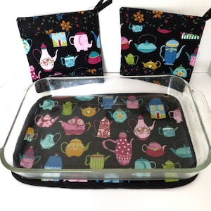 May include: Set of three black fabric pot holders with a teapot and house pattern. The pot holders are quilted and have a loop for hanging. The pot holders are on top of a glass baking dish with a matching fabric liner.