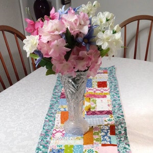 May include: A clear glass vase with a textured design holds a bouquet of pink, white, and blue flowers. The vase is sitting on a colorful patchwork table runner.