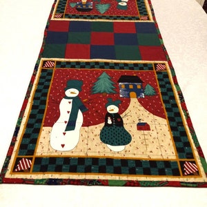 May include: A red, green, and white quilted table runner with a winter theme. The runner features a checkered pattern and a design of snowmen, trees, and houses.