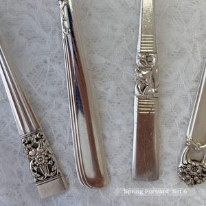 May include: Close-up of four silver-colored vintage silverware pieces. The handles feature floral and geometric designs. The text "Spring Forward Set 6" is visible in the bottom right corner. The silverware is arranged on a white, textured surface.