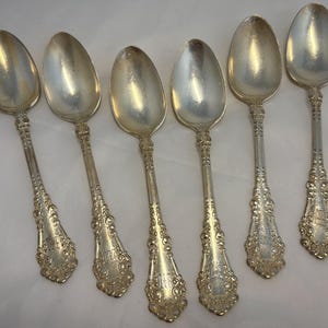 May include: Six antique silver spoons with ornate handles and oval bowls. The spoons have a reflective surface and detailed floral and scrollwork patterns on the handles. The spoons are arranged in a row, showcasing their elegant design.