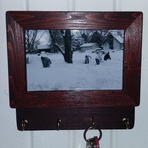 May include: A dark brown wooden key holder with a picture frame and three hooks. The picture frame contains a photo of three people shoveling snow in a snowy yard.