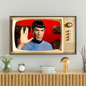 Puede incluir: Un televisor vintage con acabado de madera de grano muestra una imagen en blanco y negro de un hombre con una camisa azul haciendo el saludo vulcano. El televisor está en un estante de madera con una lámpara dorada y un jarrón de flores blancas.