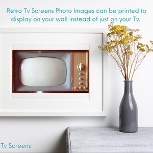 May include: A white picture frame with a vintage television screen inside. The television is brown with a white screen. The frame is hanging on a white wall. A vase with dried yellow flowers is on a white table below the frame.