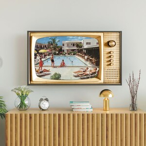 Op de afbeelding: Een ingelijste televisie in vintage-stijl toont een retro zwembadsc&egrave;ne. De tv heeft een goud en zwart frame, met een klok en knoppen aan de zijkant. De tv is gemonteerd op een witte muur boven een houten kast.