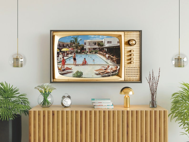 Op de afbeelding: Een ingelijste televisie in vintage-stijl toont een retro zwembadsc&egrave;ne. De tv heeft een goud en zwart frame, met een klok en knoppen aan de zijkant. De tv is gemonteerd op een witte muur boven een houten kast.