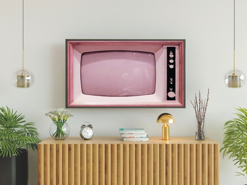 May include: A vintage pink television set with a black frame is mounted on a white wall above a wooden shelf. The shelf has a gold lamp, a clock, a vase of white flowers, and a stack of books.