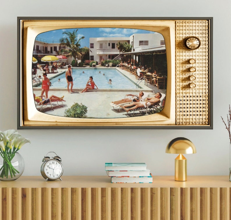 Op de afbeelding: Een ingelijste print met een vintage televisie. Het scherm toont een zonnige zwembadsc&egrave;ne met mensen die ontspannen en zwemmen. De tv-frame is goudkleurig, met een klok en bedieningsknoppen aan de zijkant.