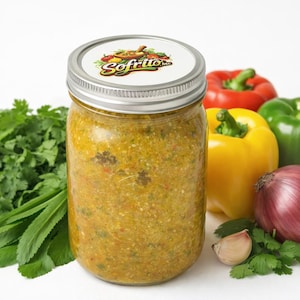 May include: A jar of sofrito salsa with a silver lid, surrounded by fresh ingredients. Red, yellow, and green bell peppers, a red onion, cilantro, and garlic are arranged around the jar. The jar's lid has the sofrito logo.