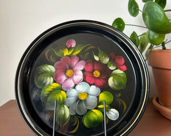 May include: A round black tray with a hand-painted floral design. The flowers are in shades of pink, red, and blue with yellow centers and green leaves. The tray has a gold trim and is displayed on a black metal stand.