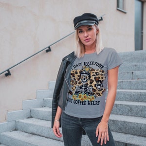 May include: Grey t-shirt featuring a skeleton wearing a leopard print headband and sunglasses holding a cup of coffee. The text on the shirt reads "I hate everyone but coffee helps."