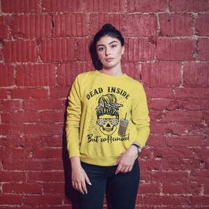 May include: Yellow sweatshirt with a skull graphic and the text "Dead inside but caffeinated". The skull is wearing sunglasses and a leopard print headband.