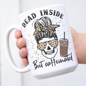 May include: White ceramic coffee mug featuring a black and white illustration of a skull wearing sunglasses and a leopard print bandana. The text "Dead inside but caffeinated" is printed on the mug.