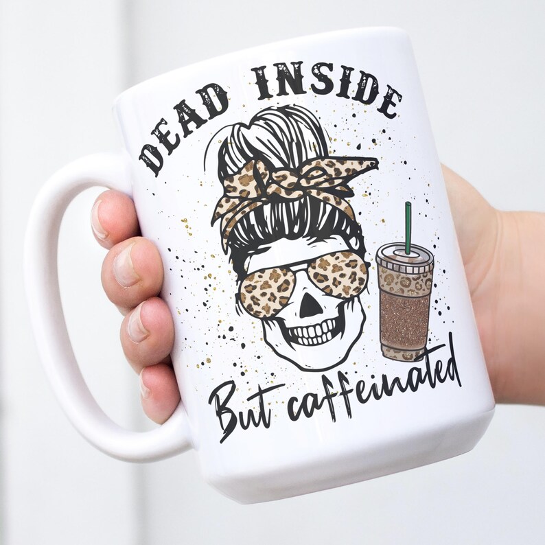 May include: White ceramic coffee mug featuring a black and white illustration of a skull wearing sunglasses and a leopard print bandana. The text "Dead inside but caffeinated" is printed on the mug.