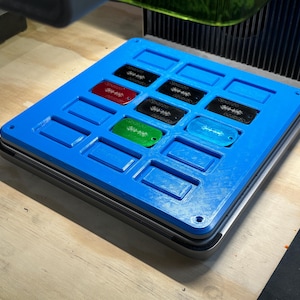 May include: A blue plastic mould with 12 rectangular slots, designed to hold small rectangular objects. The mould is sitting on a silver surface. There are 6 colourful rectangular objects in the slots, including red, green, blue, and black.