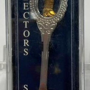 May include: A silver collector's spoon in a clear plastic case. The spoon features a decorative handle with a shield-shaped emblem and a small gold-colored charm. The spoon's bowl has an engraved design. The text "COLLECTORS SPOONS" is visible.