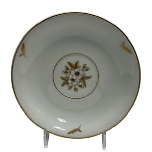 Puede incluir: Plato de cerámica blanca con borde dorado y diseño floral. El centro presenta un círculo dorado con un motivo floral de flores blancas y centros morados. Acentos de hoja de oro se colocan alrededor del borde del plato.