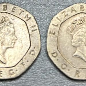 Może przedstawiać: Dwie srebrne brytyjskie monety dwudziestopensowe. Każda moneta przedstawia profil królowej Elżbiety II w koronie, otoczony tekstem. Monety leżą na szarej powierzchni.