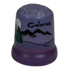 May include: A purple thimble with a landscape design. The thimble features a mountain range, a full moon, and a green tree. The word "Colorado" is written in black script on the thimble.