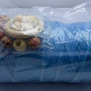 Puede incluir: Un paquete que contiene un gran rollo de hilo azul claro, con una colección de cuentas de madera y un anillo de madera. Las cuentas son en tonos marrones y beige. El hilo está bien enrollado y visible a través del embalaje de plástico transparente.