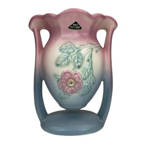 Puede incluir: Un jarrón de Hull Pottery con un diseño floral. El jarrón presenta un diseño de dos asas con una base azul claro que se desvanece en un cuerpo blanco y rosa. Una flor rosa y hojas verdes están en relieve en la parte delantera. La etiqueta Hull Pottery es visible.