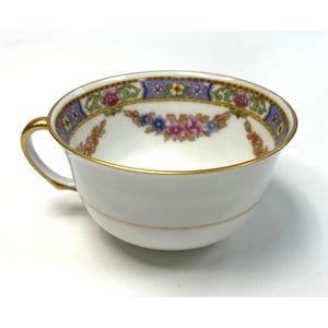 May include: White teacup with a gold handle and rim. The cup features a floral design with pink, blue, and yellow flowers, and a blue and purple border. The teacup is likely made of porcelain.