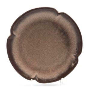 May include: A round, brown ceramic plate with a unique scalloped edge. The plate has a speckled, metallic finish, with a darker brown rim and a lighter brown center. The plate is displayed on a clear stand.
