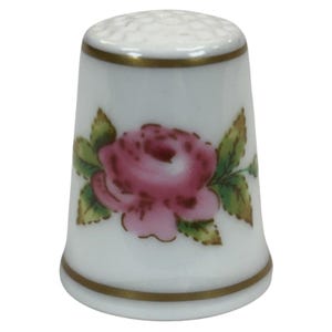 May include: White porcelain thimble with a pink rose and green leaves. The thimble has a gold rim and a textured top. A decorative sewing accessory.