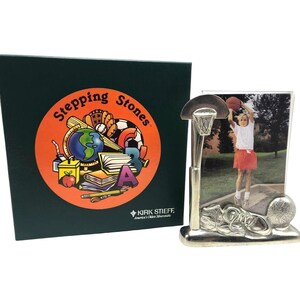 Puede incluir: Un marco de fotos plateado con temática de baloncesto, con una canasta, un balón y zapatillas deportivas. El marco contiene una foto de una niña jugando al baloncesto. Una caja verde con un logotipo circular que dice "Stepping Stones" está al lado del marco.