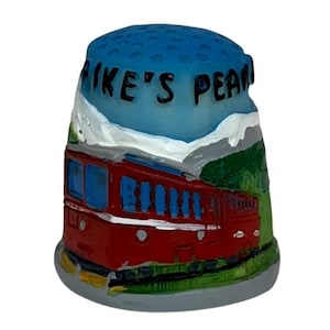 May include: A ceramic thimble featuring a red train with blue windows, set against a backdrop of green hills and white clouds. The thimble has a blue top with the text "PIKE'S PEAK" in black.