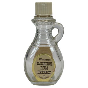 Puede incluir: Una pequeña botella de vidrio transparente con asa y tapón beige. La etiqueta dice "Watkins IMITATION RUM EXTRACT 2 FLUID OZ". La botella tiene un diseño decorativo alrededor del cuello.
