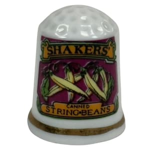 Può includere: Un ditale in porcellana bianca con una fascia dorata alla base. Il ditale presenta un'etichetta con il testo "SHAKERS" e "CANNED STRING BEANS" insieme a un'illustrazione di fagiolini.