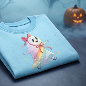 Könnte beinhalten: Hellblaues Sweatshirt mit einem Cartoon-Geist-Design. Der Geist hat einen Regenbogenkörper, eine rosa Schleife und lächelt. Ein geschnitzter Kürbis ist im Hintergrund zu sehen, was auf ein Halloween-Thema hindeutet.