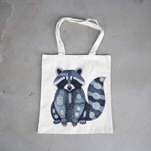 Könnte beinhalten: Weißer Shopper mit einem Waschbär-Design. Der Waschbär ist aus blauem Denim mit Used-Details gefertigt. Die Tasche hat zwei weiße Henkel und ist etwa 40 cm hoch.