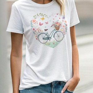 Könnte beinhalten: Weißes T-Shirt mit einem floralen Herzdesign, das ein Fahrrad mit einem Blumenkorb zeigt. Schmetterlinge und Herzen sind um das Design verstreut.