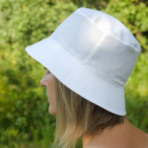 Può includere: Un cappello a secchiello bianco con un'ampia tesa. Il cappello è fatto di un tessuto morbido e leggero.