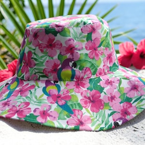 Op de afbeelding: Een bucket hat met een levendige tropische print van roze hibiscusbloemen, groene palmbladeren en kleurrijke papegaaien op een witte achtergrond. De hoed heeft een brede, naar beneden hellende rand en een platte bovenkant met bloemen.