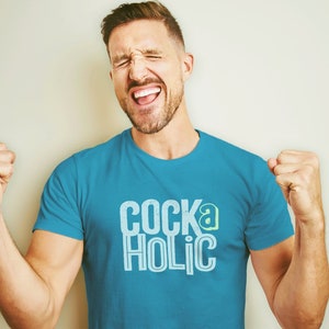 May include: A man wearing a turquoise blue t-shirt with the text "COCKaHOLIC" in white and green letters. He is smiling and has his fists clenched in the air.