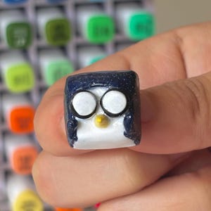May include: A small, square, penguin-shaped artisan keycap. The keycap is primarily navy blue with a white face, black eyes, and a yellow beak. The keycap is held in front of a background of colorful markers.