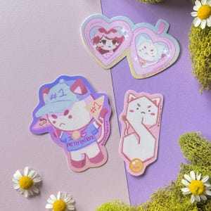 May include: A collection of colorful stickers featuring cartoon cat characters. One sticker says "#1 Pretty Patrick". Other stickers include heart-shaped designs with cartoon characters. Daisies and green moss are also present.