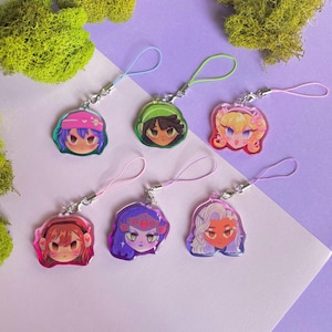 May include: Six colorful acrylic charms featuring stylized anime-style faces. Each charm has a different hair color and accessory, with a matching colored loop and silver-tone clasp. The charms are displayed on a purple and white surface.